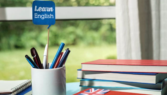 Escritorio con material de estudio para aprender inglés, incluyendo un vaso con bolígrafos de colores y libros apilados, con una bandera del Reino Unido visible.