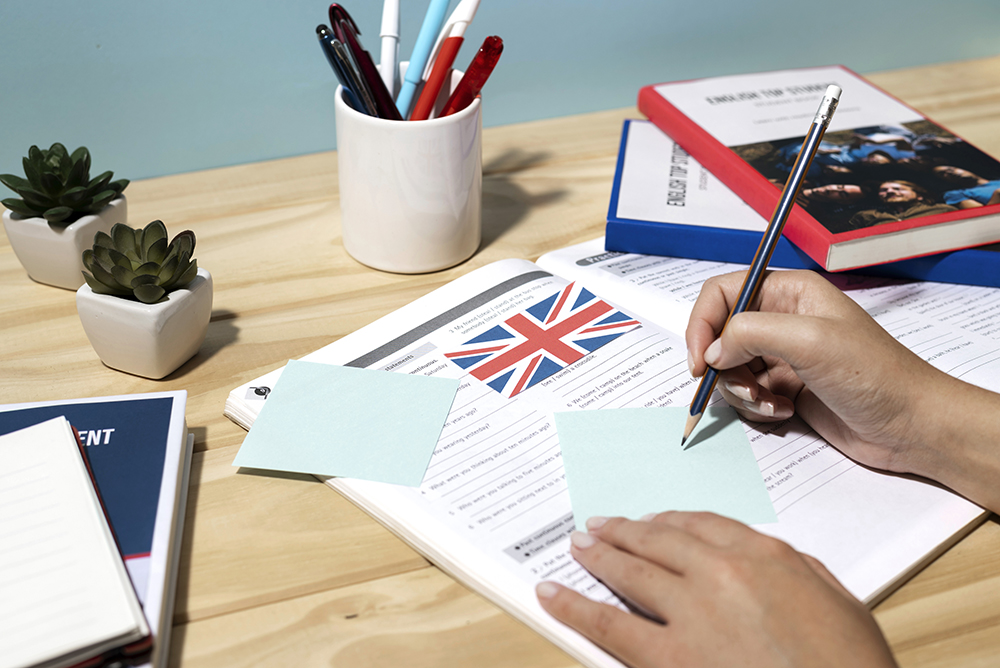 Mano escribiendo en una libreta con una bandera del Reino Unido, rodeada de lápices, macetas con plantas y libros sobre el aprendizaje del inglés, en un ambiente de estudio.