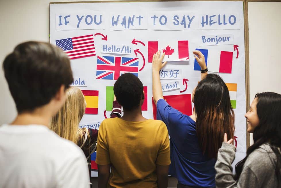 Grupo de estudiantes interactuando con un cartel que presenta diferentes formas de decir "hola" en varios idiomas, incluyendo inglés, español y francés, mientras colocan banderas en un mural educativo.