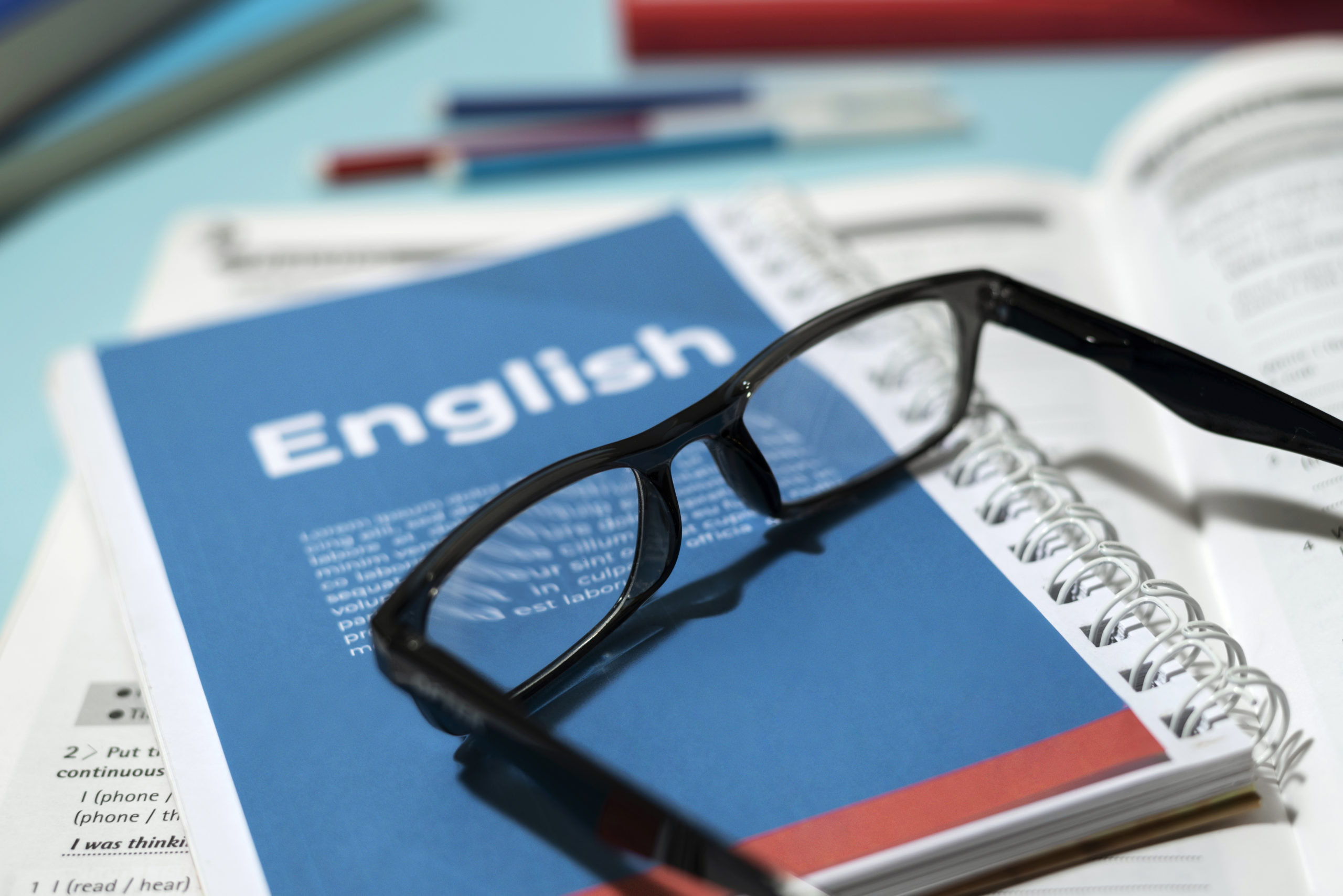 Gafas sobre un libro de inglés con una portada azul, rodeado de materiales de estudio como cuadernos y bolígrafos, en un entorno de aprendizaje.