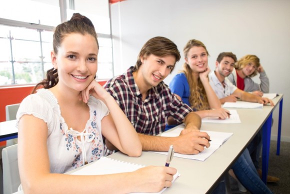Grupo de estudiantes sonrientes en un aula, sentados en escritorios con libretas y bolígrafos, listos para tomar notas. La imagen muestra un ambiente de aprendizaje colaborativo y positivo.